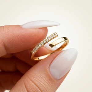 Op de afbeelding: Een open gouden ring met een gepolijste band, waarbij het ene uiteinde is versierd met een rij kleine, heldere, sprankelende steentjes en het andere uiteinde een gladde, platte, goudkleurige afwerking heeft voor een verfijnd sieraad.