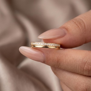 May include: A gold ring featuring a thin band with a row of small, clear, sparkling stones embedded along the top half. The ring has a polished, smooth finish on the lower half of the band, creating a refined and classic appearance for this jewelry.
