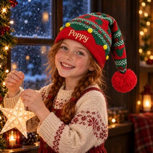 May include: A red and green knit elf hat featuring a folded brim with the name Poppy embroidered in white thread, small yellow pom-poms along the edge, and a large red yarn pom-pom at the pointed tip with a patterned design.