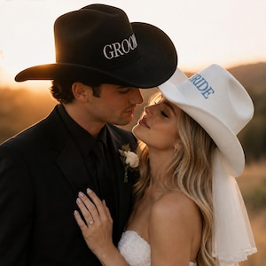Op de afbeelding: Een zwarte cowboyhoed met het woord GROOM in witte letters en een witte cowboyhoed met het woord BRIDE in blauwe letters, voorzien van een korte witte sluier aan de achterkant van de witte hoed, beide gedragen in een buitenomgeving.