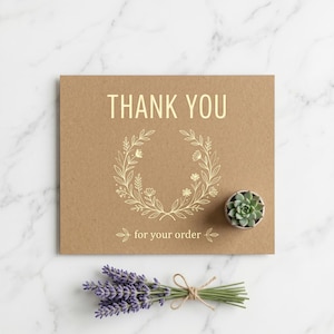 Peut inclure: Une carte en papier kraft avec les mots "THANK YOU" et "for your order" en lettres dor&eacute;es, ainsi qu'un motif de couronne florale. Une petite plante succulente dans un pot gris et un bouquet de lavande sont &eacute;galement pr&eacute;sents.