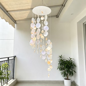 Op de afbeelding: Een hangende mobiel met talloze ronde, iriserende capiz-schelpschijven die met dunne draden aan een wit rond frame hangen. De doorschijnende schelpen hebben een parelmoerglans met zachte beige en witte tinten, gerangschikt in een trapsgewijs spiraalpatroon dat een gelaagd effect creëert.