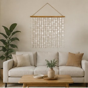 Puede incluir: Un tapiz de pared compuesto por múltiples hilos verticales de discos de concha nácar blanca translúcida suspendidos de una varilla de madera horizontal con un cordel fino para colgar, creando un efecto de cortina superpuesta y semitransparente.