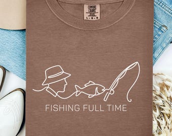 Puede incluir: Una camiseta marrón Comfort Colors con un dibujo lineal blanco de una persona con sombrero, un pez y una caña de pescar. El texto "FISHING FULL TIME" está debajo. La camiseta está rodeada de mezclilla y un sombrero.