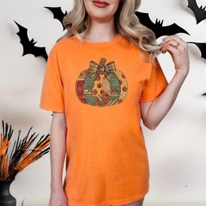May include: An orange short-sleeve t-shirt featuring a central graphic of a pumpkin. The pumpkin is composed of various patterned segments including apples, alphabet letters, and math symbols, topped with a plaid bow in shades of green and brown.