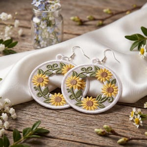 Peut inclure: Une paire de boucles d'oreilles pendantes circulaires en tissu blanc brodé de tournesols jaunes, de cœurs marron et de feuilles vertes. Les boucles d'oreilles sont dotées de crochets en métal argenté et d'un cadre circulaire mettant en valeur la broderie.