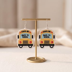 May include: A pair of embroidered school bus earrings featuring a yellow bus body, black bumpers, red lights, and a light blue windshield with the black text SCHOOL BUS and BACK TO SCHOOL stitched onto the front of each earring piece.