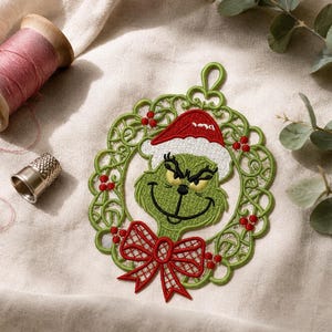 May include: A green embroidered character ornament featuring a red Santa hat and a red bow, framed by a circular green wreath with red berries. The ornament includes a top loop for hanging and is displayed alongside a wooden spool of pink thread.