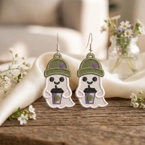 May include: A pair of dangle earrings featuring embroidered white ghosts wearing olive green beanies with small purple hearts. Each ghost holds a purple coffee cup with a black lid and straw, suspended from silver-toned metal earring hooks.
