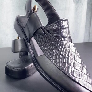 Pode incluir: Sandálias de couro preto com um padrão texturizado semelhante ao de um crocodilo na parte superior. As sandálias têm uma tira no tornozelo e um fecho de fivela. A sola é plana e o design é elegante e moderno.
