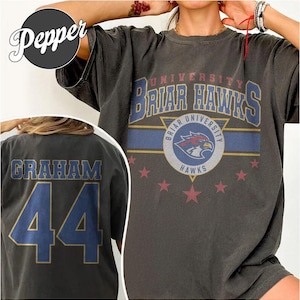 Può includere: T-shirt grigio scuro dell'Università Briar Hawks. La parte anteriore presenta la scritta "Briar Hawks University" in blu e oro, con un logo di falco e stelle rosse. Il retro mostra "GRAHAM 44" in blu e oro.