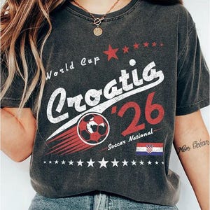 May include: A charcoal gray t-shirt featuring a distressed graphic print that reads World Cup Croatia '26 Soccer National. The design includes a red soccer ball, a row of stars, and the Croatian flag in red, white, and blue colors.