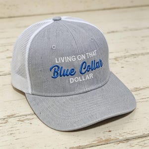 Puede incluir: Gorra de camionero gris con parte trasera de malla blanca y visera curva. La gorra presenta el texto bordado "LIVING ON THAT Blue Collar DOLLAR" en hilo azul. Adecuada para uso informal.