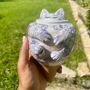 Peut inclure: Une sculpture en céramique en forme de chat, dans des tons de blanc et de bleu, tenue dans une main. La sculpture présente des motifs tourbillonnants complexes et des détails de pattes. La base est en forme de cône. L'arrière-plan est une pelouse verte floue.