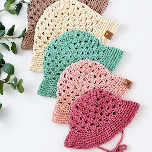 Puede incluir: Una colección de cinco sombreros de pescador de ganchillo en marrón, crema, verde azulado, rosa empolvado y rosa oscuro. Cada sombrero presenta un patrón texturizado de punto abierto y una pequeña etiqueta de cuero marrón.