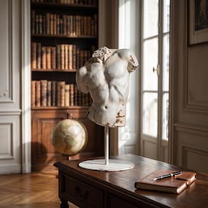 May include: A white, weathered sculpture of a male torso on a metal stand. The sculpture is displayed on a dark wooden table, with a leather-bound notebook and pen. A globe and a bookcase filled with books are in the background.