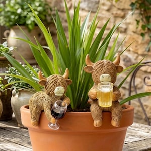 May include: Two brown Highland cow figurines perched on the rim of a terracotta plant pot. The left cow holds a miniature wine glass with red liquid, while the right cow holds a miniature beer mug filled with a golden liquid and white foam.