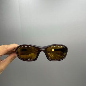May include: Dark brown sunglasses featuring a wrap-around frame design with a series of small cutouts along the upper and lower edges of the lenses. The lenses are tinted a deep amber color, and the word SATISFY is printed in white on the bridge.
