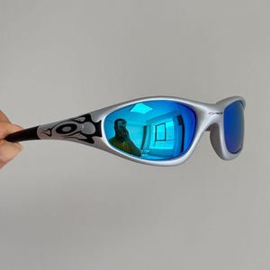 Può includere: Occhiali da sole color argento con montatura avvolgente e lenti a specchio blu. Le aste presentano un motivo grafico nero di ispirazione tribale e il logo Oakley è visibile sull'angolo superiore della montatura della lente destra.