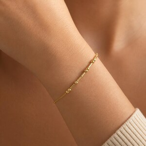 May include: A delicate gold chain bracelet featuring a series of small, polished gold beads and short cylindrical bars arranged in a linear pattern along the centre, creating a subtle, minimalist design that rests against the skin.
