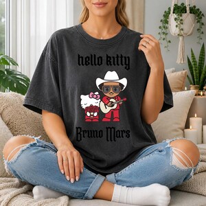 May include: Dark gray t-shirt featuring a graphic of Hello Kitty and Bruno Mars. Hello Kitty is holding flowers, and Bruno Mars is wearing a cowboy hat and playing a guitar. The text "hello kitty" and "Bruno Mars" are printed on the shirt.