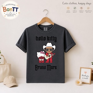 May include: Dark gray t-shirt featuring a graphic of Hello Kitty and Bruno Mars. The design includes the text "hello kitty" above the image and "Bruno Mars" below. The shirt is hanging on a wooden hanger.