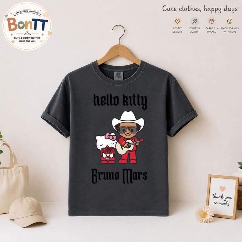 May include: Dark gray t-shirt featuring a graphic of Hello Kitty and Bruno Mars. The design includes the text "hello kitty" above the image and "Bruno Mars" below. The shirt is hanging on a wooden hanger.