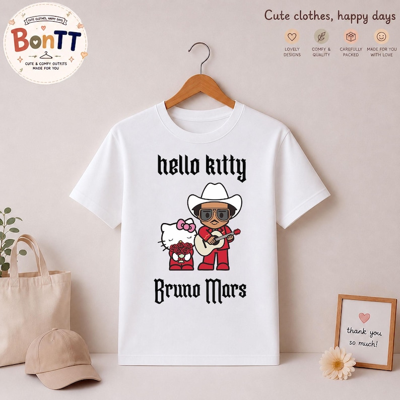 May include: White t-shirt featuring a graphic of Hello Kitty and Bruno Mars in a cowboy hat playing a guitar. The text "hello kitty" and "Bruno Mars" are printed in black gothic font. The shirt is hanging on a wooden hanger.
