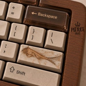 Puede incluir: Un teclado mecánico con marco de madera oscura y teclas beige. Una tecla de retroceso marrón translúcida tiene el texto Backspace. La tecla enter muestra un gráfico de pez fosilizado marrón. El texto PIERCE PLUG con una corona es visible.