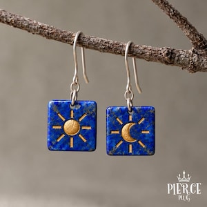 Puede incluir: Un par de pendientes cuadrados colgantes de piedra lapislázuli azul intenso con incrustaciones doradas. Un pendiente muestra un motivo de sol con rayos, mientras que el otro presenta un diseño de media luna. Ambos cuelgan de ganchos de metal plateado.