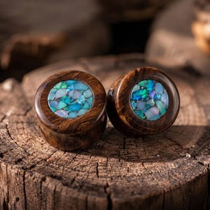 Könnte beinhalten: Ein Paar kreisförmige Ohrplugs aus dunkelbraunem Holz mit polierter Oberfläche. Die Mitte jedes Plugs ist mit einer Einlage aus zerkleinerten, schillernden Opalsteinen in Blau-, Grün- und Weißtönen versehen, die ein Mosaikmuster bilden.