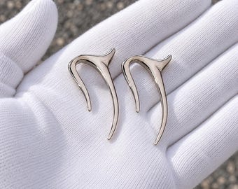 Puede incluir: Un par de pendientes de presión de metal plateado con un diseño liso, curvado y puntiagudo. Las piezas de joyería tienen un acabado pulido y reflectante, con una forma elegante y alargada que sigue un arco sutil sobre tela blanca.