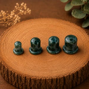 May include: Four smooth, polished, dark green stone cups with rounded tops and flared bases, arranged in a row from smallest to largest. Each cup features natural mottled patterns in shades of deep forest green and black throughout the stone surface.