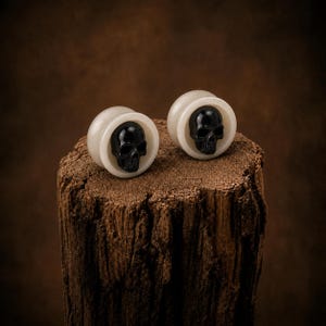 May include: A pair of circular ear plugs featuring a smooth, off-white outer rim and a detailed, glossy black skull centered within each piece. The design showcases a high-contrast aesthetic with a polished finish on the skull and a matte texture on the surrounding ring, creating a distinct visual focus for these body jewelry items.