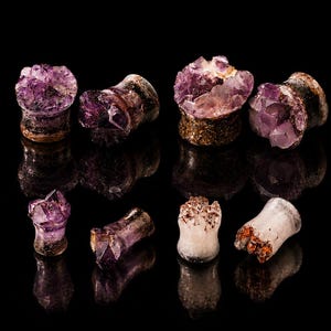 Puede incluir: Colección de expansores de piedra amatista en varias formas y tamaños. Los expansores presentan un tono púrpura intenso con estructuras cristalinas, sobre una superficie negra reflectante. Joyería corporal con estética de piedras preciosas.