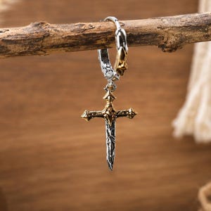 Puede incluir: Un pendiente de aro en tonos plata y oro con un colgante en forma de espada con guarda en cruz. El metal tiene un acabado texturizado y desgastado con detalles en oro pulido en la guarda y en la parte superior del aro.