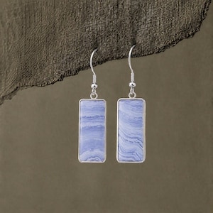 May include: A pair of dangle earrings featuring rectangular blue lace agate stones set in silver-toned metal frames. Each earring hangs from a simple silver-toned hook wire, showcasing the natural light blue and white banded patterns of the stone.