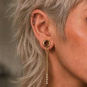 May include: A gold-toned ear tunnel gauge featuring a dangling chain attachment. The jewelry includes a circular tunnel plug, a short vertical chain segment with three small rings, and a long thin chain ending in four small clear round stones.