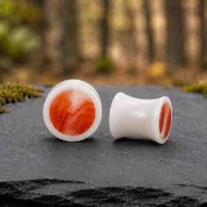 May include: A pair of ear plugs featuring a smooth white outer rim and a vibrant orange marbled center. The plugs have a concave, flared shape, with one piece shown from the front and the other positioned to display its side profile and depth.