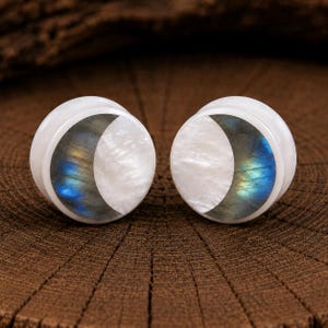 Pode incluir: Um par de alargadores circulares com um design de meia-lua. Cada peça é feita com uma incrustação de madrepérola branca combinada com uma secção de pedra labradorite cinzenta que exibe reflexos de luz azul e amarela.