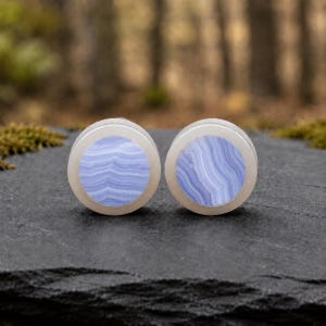 May include: A pair of circular ear plugs featuring a smooth, translucent white outer rim and a central inlay of blue lace agate stone. The stone displays distinct, wavy, light blue and white banded patterns across the flat, circular surface.