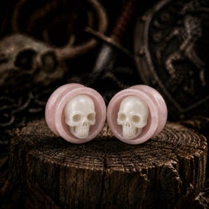 May include: A pair of circular ear plugs featuring a smooth, pale pink stone rim and a carved, off-white skull centered within each piece. The plugs have a polished finish and a distinct cylindrical shape, showcasing the contrast between the soft pink stone and the detailed, light-colored skull carvings.