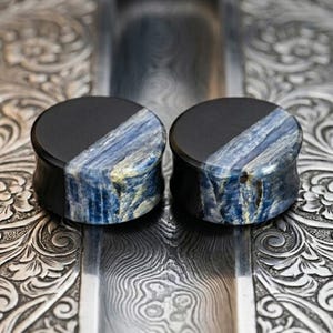 Op de afbeelding: Twee zwarte en blauwe stenen oordopjes. De oordopjes hebben een zwarte bovenkant en een diagonale sectie van blauwe en witte steen. Ze worden gepresenteerd op een zilveren, bewerkte ondergrond.