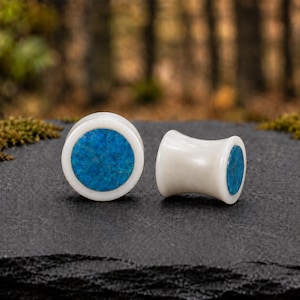 May include: A pair of circular ear plugs featuring a smooth white body with a vibrant blue stone inlay on the front face. The plugs have a concave, flared shape, with one piece shown from the front and the other positioned at a side angle.