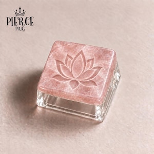 May include: A square keycap featuring a polished pink stone top with an engraved lotus flower design. The base of the keycap is made of clear, transparent material. The text PIERCE PLUG with a crown icon is visible in the upper left corner.