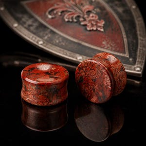 May include: Two red jasper stone ear plugs with a dark, mottled pattern. The plugs are cylindrical with a flared edge. The background features a decorative shield with a fleur-de-lis design.