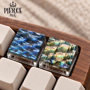 May include: Two square artisan keycaps with a textured, wavy surface made of labradorite stone. One keycap displays deep blue iridescent tones, while the other features golden-green hues. They are set into a wooden keyboard frame with white keys.
