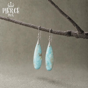 Puede incluir: Un par de pendientes colgantes con piedras de color azul claro en forma de lágrima alargada con vetas blancas y marrones. Las piedras están engastadas en tapas de metal plateado y cuelgan de ganchos plateados. Texto PIERCE PLUG visible.