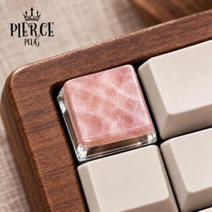 May include: A square mechanical keyboard keycap featuring a polished, translucent pink stone top with a natural crystalline texture, set within a dark wood keyboard frame alongside several plain white keycaps. The text PIERCE PLUG is visible.