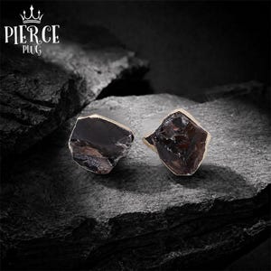 May include: A pair of stud earrings featuring raw, dark brown, translucent stones with irregular, jagged edges and metallic gold-toned settings. The text PIERCE PLUG with a crown icon is visible in the upper left corner of the image.